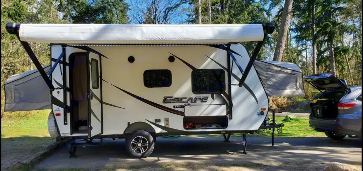 White Escape travel trailer rv rental set up at a wooded campsite, awning out, SUV trunk open for gear.. K-Z Manufacturing Escape 2017