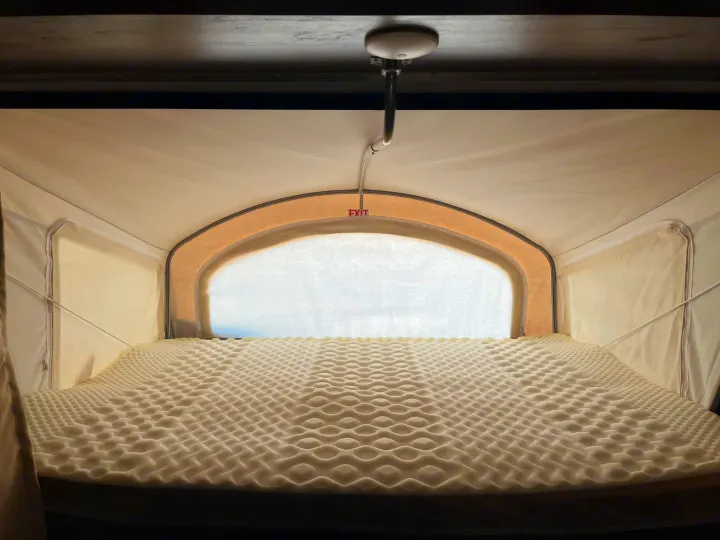 Interior view of an RV rental pop-up camper bunk, featuring a textured foam mattress topper and an overhead exit sign.. K-Z Manufacturing Escape 2017