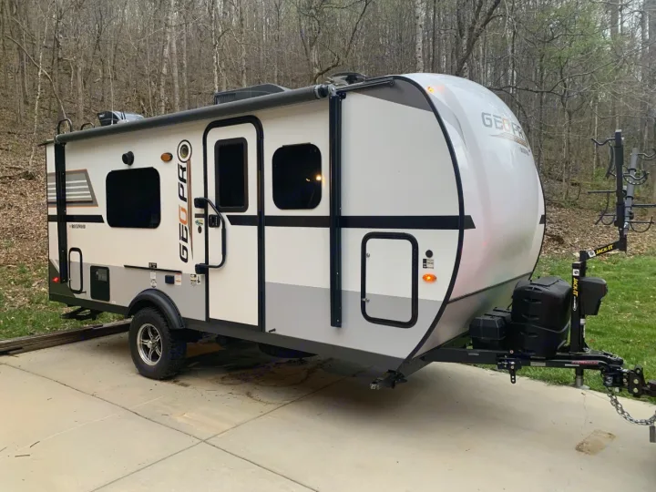 Modern white and gray Geo Pro travel trailer, ideal for an RV rental or camping trip. Parked on concrete with a wooded backdrop.. Forest River Rockwood 2020