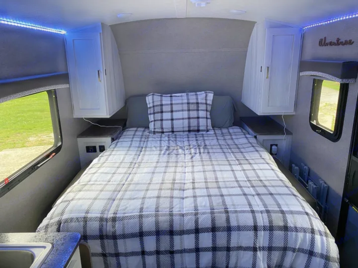 Inside a cozy RV rental bedroom: a plaid bed, overhead cabinets, and soft blue lighting create a comfortable space.. Forest River Rockwood 2020