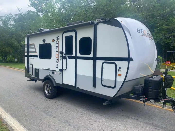 This white & gray Geo Pro travel trailer, perfect as an RV rental, awaits adventure on a paved road surrounded by trees.. Forest River Rockwood 2020