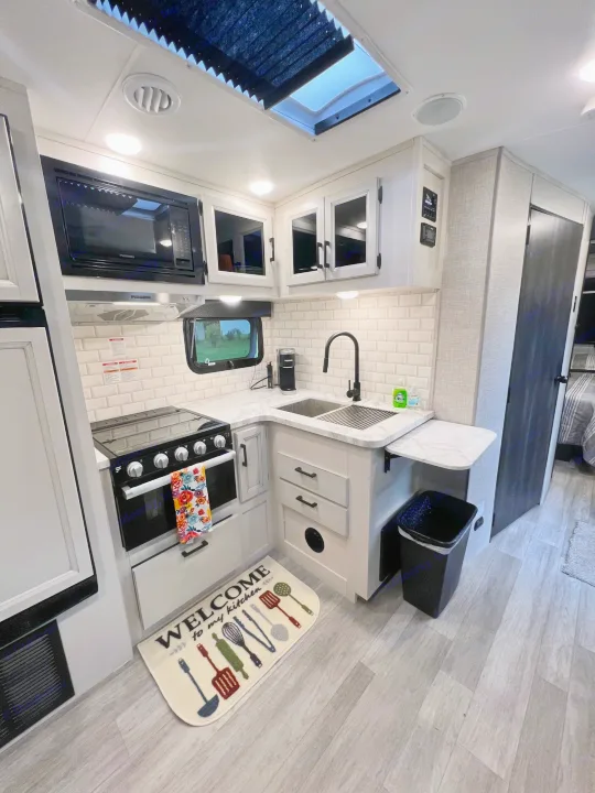 Step into this bright RV rental kitchen, featuring subway tiles, stove, microwave, sink, and a cozy 'Welcome' mat.. Jayco White Hawk 2022