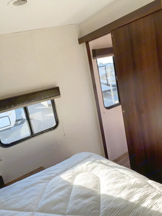 Inside an RV rental bedroom: white bedding on a bed, twin windows revealing other trailers, and a dark sliding door.. Jayco Octane 2019