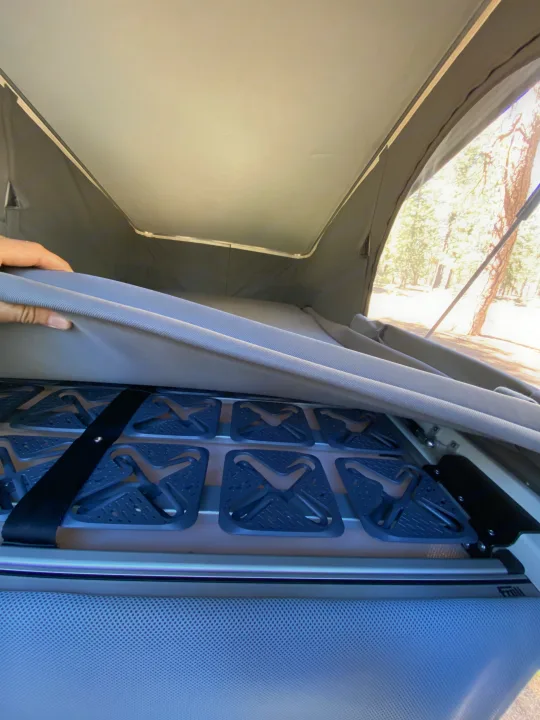 A hand lifts an RV rental mattress, revealing a Froli modular bed base for improved sleep. Pop-top tent and trees visible outside.. Mercedes-Benz Metris Weekender 2021