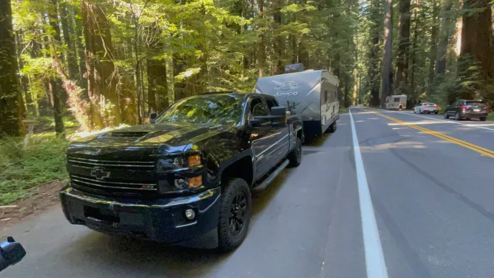 Scenic road trip with an RV rental. A black pickup tows a Jayco travel trailer through a tall forest.. Jayco Octane 2019