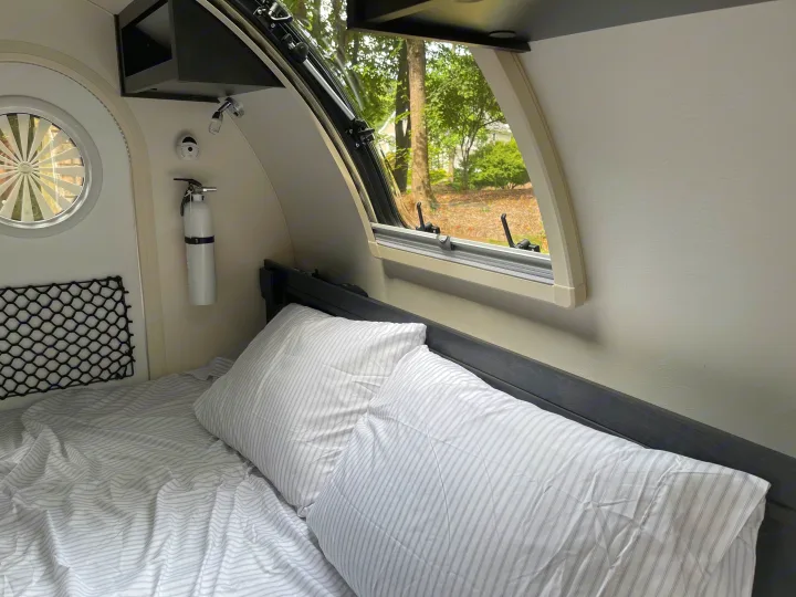Cozy RV rental interior with a bed, window view of trees, and a fire extinguisher. Ideal for comfortable travel adventures.. nuCamp RV T@G XL 6 wide 2022