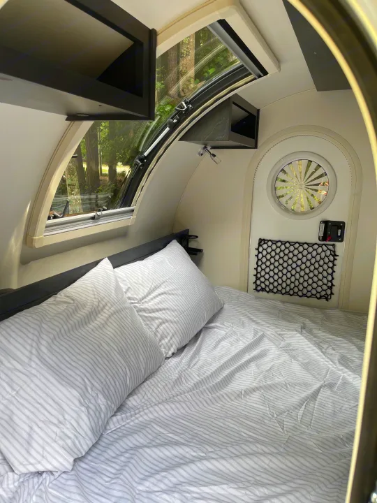 Cozy RV rental interior: a bed with striped white bedding, overhead storage, a large window revealing trees, and a compact design.. nuCamp RV T@G XL 6 wide 2022