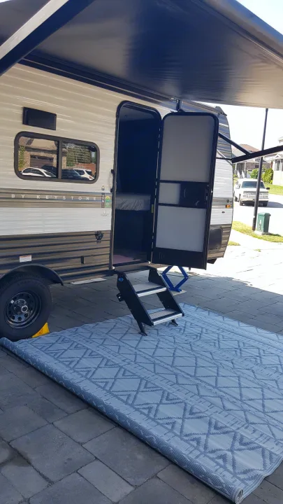 RV rental setup: open door, extended awning, portable steps, and large blue outdoor mat on a paved patio.. Keystone RV Hideout 2022