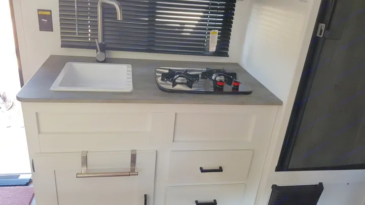 Compact RV rental kitchen features a sink, two-burner stove, white cabinets, and dark blinds on the window.. Keystone RV Hideout 2022