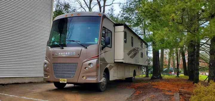 Tan Winnebago motorhome, a great RV rental option, parked at home beside a house & pine trees.. Winnebago Vista 2016
