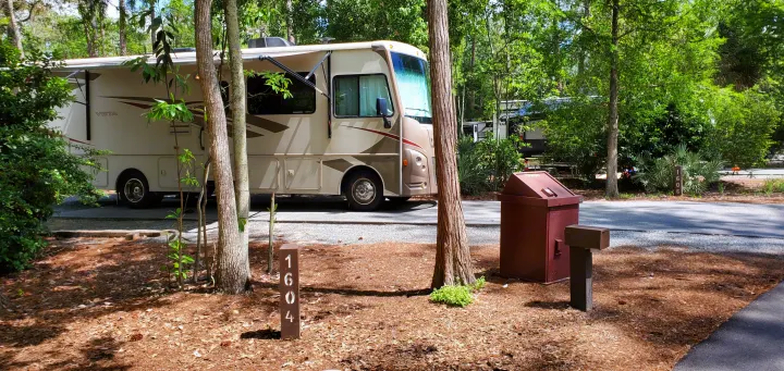 A beige RV rental motorhome parked on a sunny campground site (1604) among trees, with an extended awning.. Winnebago Vista 2016