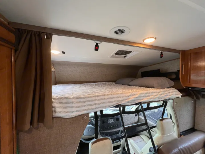 Cozy over-cab bunk bed in an RV rental, featuring striped bedding, a ladder, and overhead lighting.. Other Other 2018
