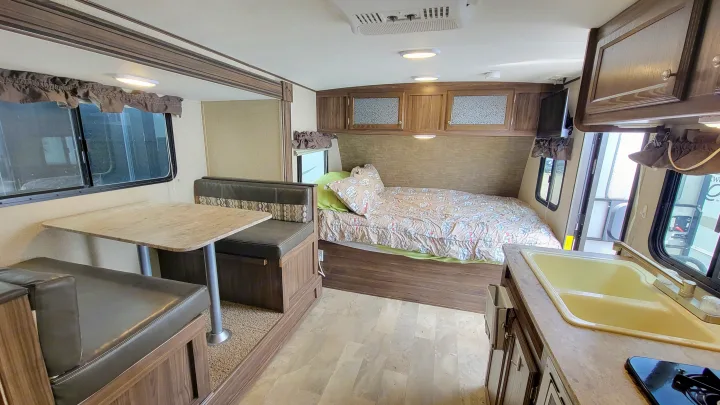 RV rental interior showing a brown dinette, bed with patterned comforter, and a kitchen area with a yellow sink.. Coachmen Apex 2018