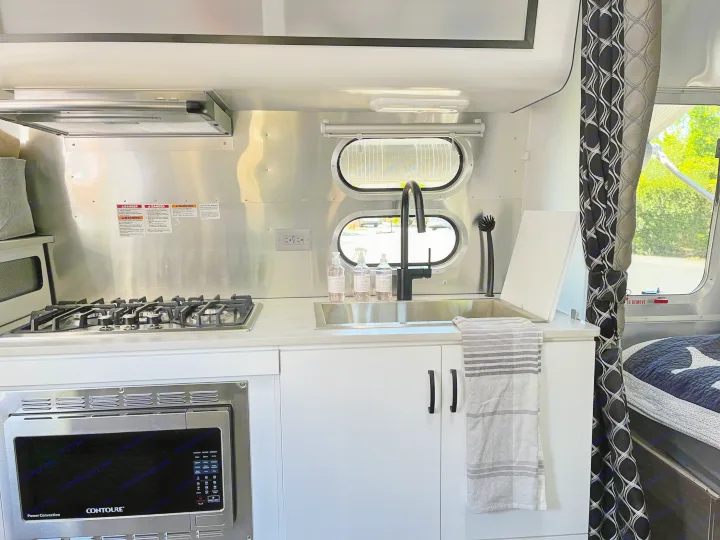 Inside a sleek RV rental, a compact kitchen features a shiny stovetop, microwave, and sink with a modern black faucet.. Airstream Caravel 2022