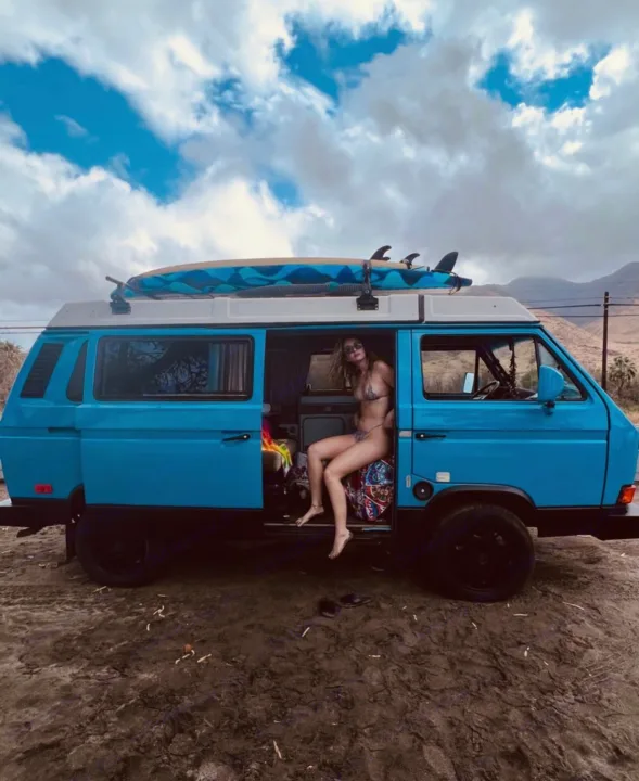 Blue camper van, perfect for a beach RV rental getaway! A woman relaxes inside, surfboard on top. Ready for adventure!. VW Westfalia 1990