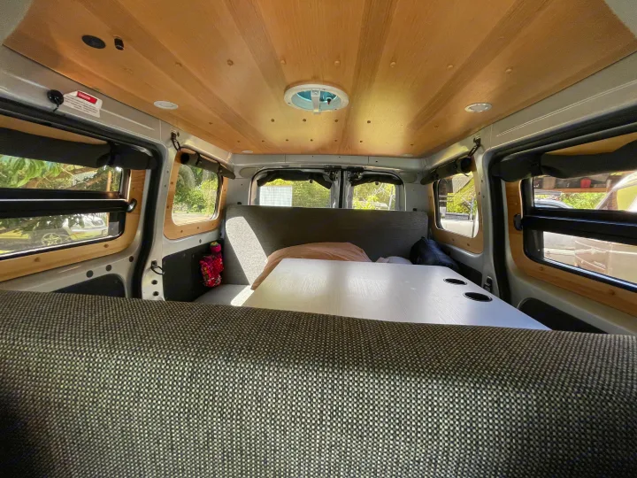 Inside an RV rental camper van: wood-paneled ceiling with fan, central table with cupholders, and upholstered seating.. Nissan NV200 2021