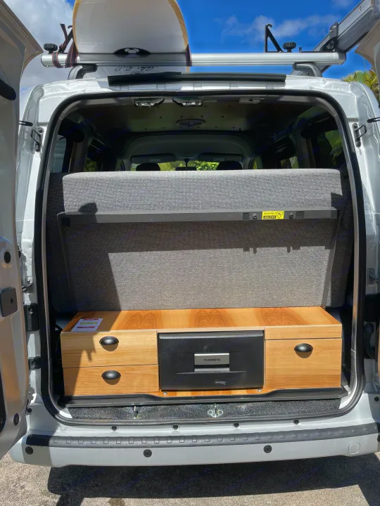 Rear view of an RV rental: Silver campervan interior with gray pull-out bed, wooden storage, Dometic fridge, and surfboard on roof rack.. Nissan NV200 2021