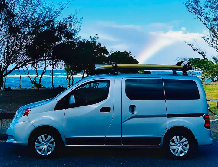 Scenic rv rental parked by the ocean with a surfboard on top & rainbow. Perfect for coastal adventures.. Nissan NV200 2021