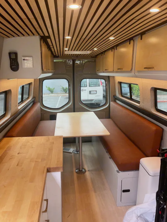 Explore this modern rv rental interior: wood slat ceiling, plush brown seating, dining tables, and bright lighting.. Mercedes Sprinter 2021