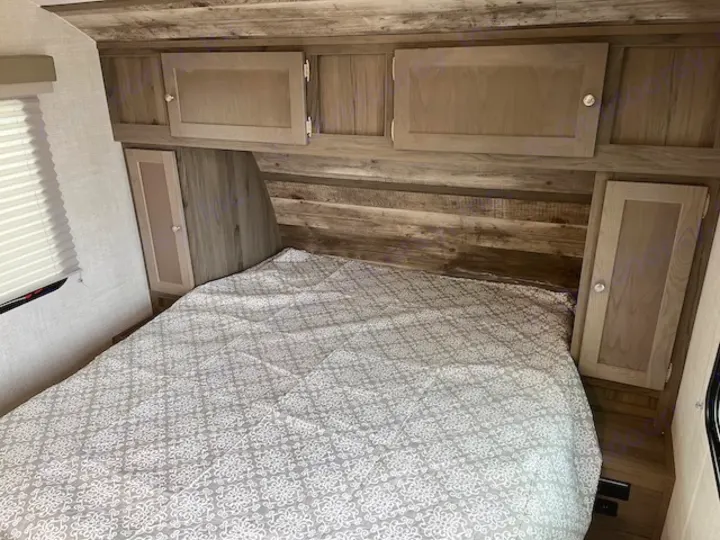 Inside an RV rental, a cozy bedroom with a patterned bed, overhead cabinets, and wood-look finishes.. Gulf Stream 21QBS 2022