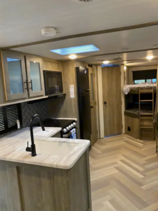 Modern RV rental interior. Spacious kitchen has a white sink, black faucet, stove, and fridge. Bunk beds & herringbone floors are visible.. Forest River Salem Hemisphere HL 2021