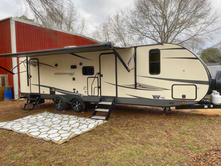 A cream-colored RV with an extended awning is parked outdoors near a red barn, ideal for an rv rental experience.. Forest River Salem Hemisphere HL 2021