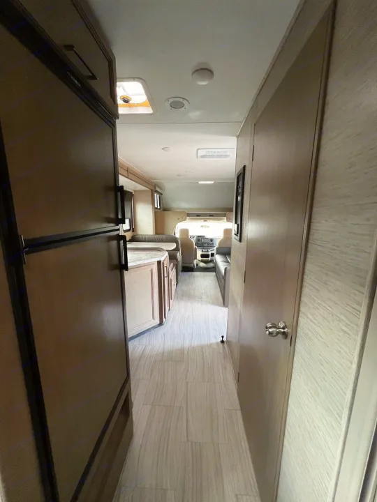 View of an RV rental's clean interior: a hallway features a large fridge on the left, a closed door on the right, and the driver's cab ahead.. Coleman Other 2022