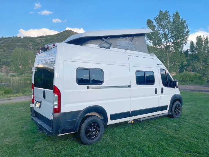 White Winnebago campervan for RV rental, parked on grass by a lake with pop-top open, ideal for nature travel.. Winnebago Solis 2021