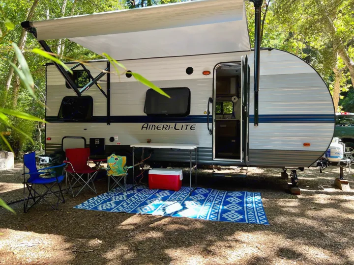 An Ameri-Lite RV rental at a shady campsite with an extended awning, outdoor chairs, table, and mat setup.. Gulf Stream Amerilite 2022