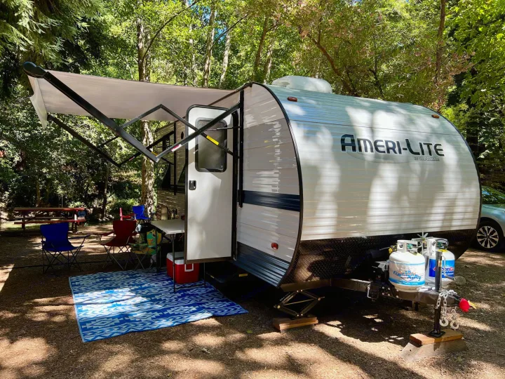 White RV rental travel trailer with an extended awning, chairs, and mat on a blue rug at a shaded, wooded campsite.. Gulf Stream Amerilite 2022