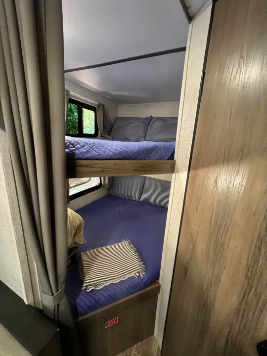 Cozy bunk beds in an RV rental, featuring blue bedding, striped pillows, and a window view. Perfect for family trips!. Gulf Stream Amerilite 2022