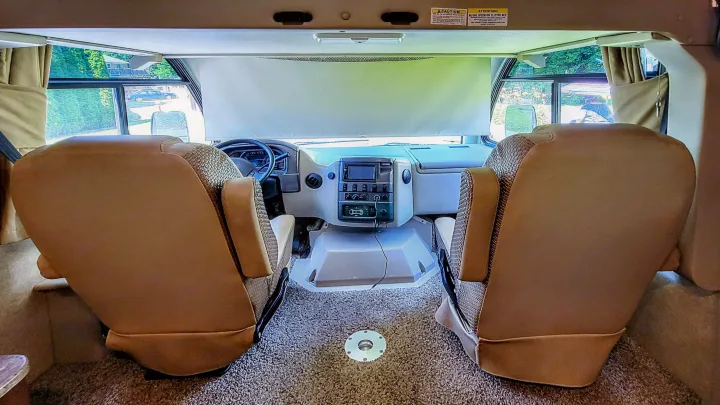 Step inside this RV rental's spacious cockpit. View from rear showing beige driver and passenger seats, dashboard, and large windshield.. Thor Vegas 2014