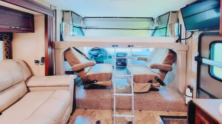 RV rental interior featuring front captain's chairs, an overhead bunk bed with ladder, a cozy sofa, and a wall-mounted TV.. Thor Vegas 2014