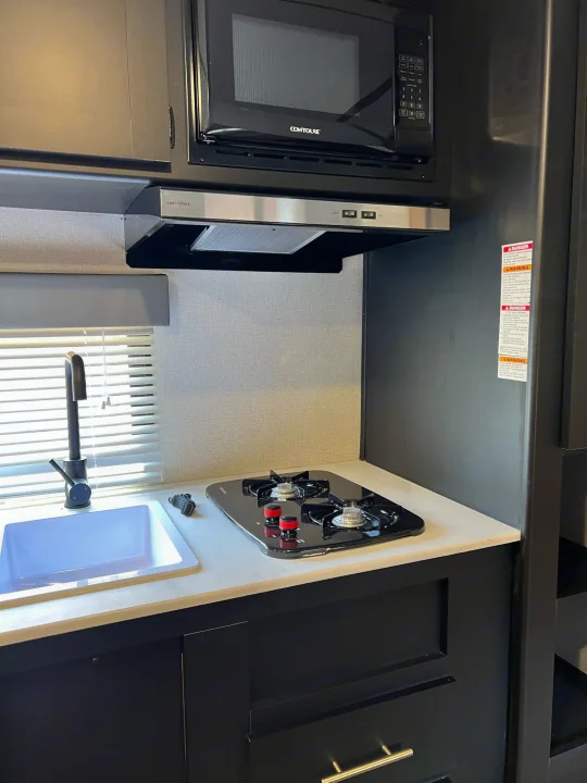 Compact kitchen in an RV rental, showing a sink, modern black faucet, two-burner stove, microwave, and dark cabinets.. Keystone Springdale 2022