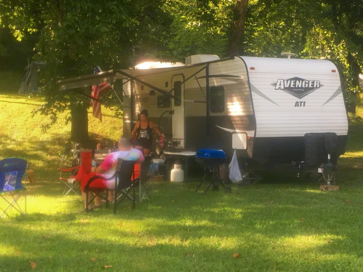 An Avenger travel trailer (RV rental) at a sunny campsite. Two people relax near chairs & a grill.. Avenger ATI 2018