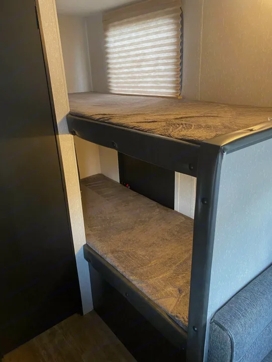 RV rental bunk beds: two compact sleeping berths with textured grey bedding, light walls, and a window with blinds.. Forest River Wolf Pup 2021