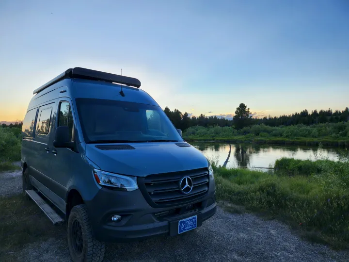 A gray Mercedes Sprinter camper van, perfect for an RV rental, is parked beside a calm river at sunset. Scenic nature view.. Winnebago Revel 2022