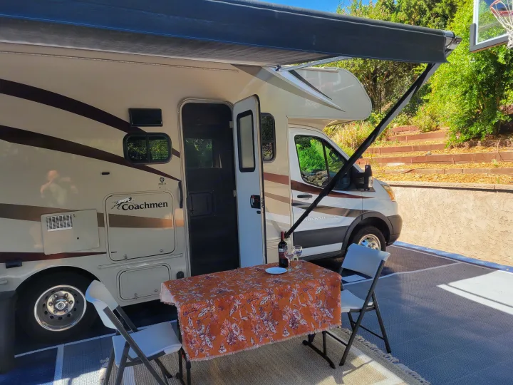 Relaxing at an RV rental campsite. A Class C motorhome with its awning extended shades a table set with wine and chairs.. Coachman Leprechaun 2018