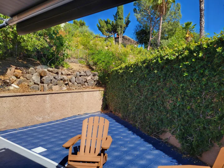 An inviting RV rental outdoor space with a blue court, Adirondack chair, stucco wall, and lush green hillside.. Coachman Leprechaun 2018
