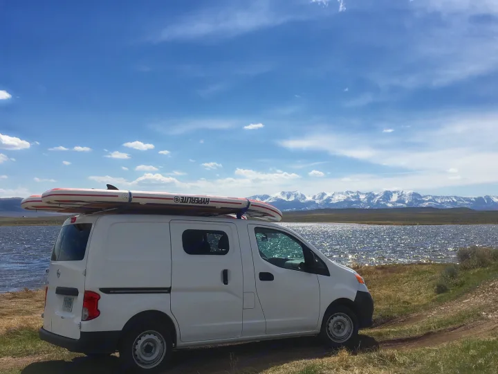 White van with paddleboards on roof by a sparkling lake, snow-capped mountains in distance. Great rv rental for adventure!. Nissan PRIMASTAR 2014