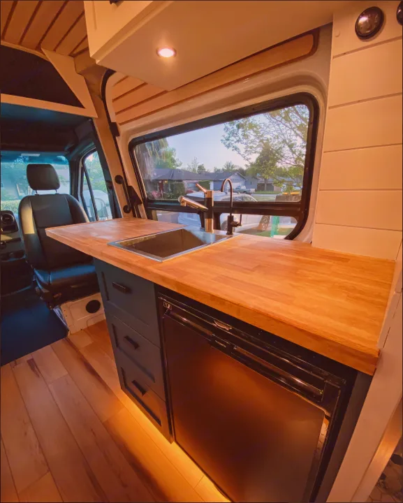 This RV rental showcases a modern camper van kitchen with a wooden countertop, sink, fridge, and sleek dark blue cabinets.. Mercedes-Benz Sprintervan 2019