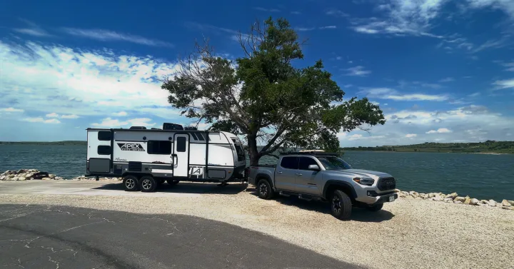 Silver pickup and Apex travel trailer parked by a lake with a tree under blue skies. Great rv rental for outdoor adventures!. Forest River Coachmen Apex Nano Off-Grid 2022