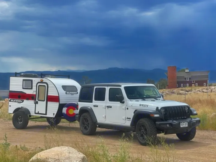 A white Jeep Wrangler tows a white SunRay RV rental, Colorado flag on spare tire. Mountains & red barn in distance.. Sunset Park RV SunRay 2020