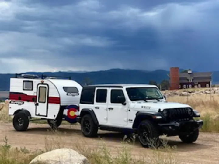 This white Jeep Wrangler tows a compact RV rental trailer, perfect for off-road camping trips in Colorado's mountains.. Sunset Park RV SunRay 2020