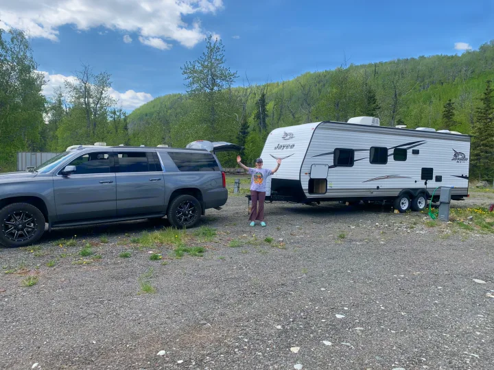 Happy with their RV rental, a person stands between a grey SUV and white travel trailer at a gravel campsite.. Jayco Jay Flight 2017
