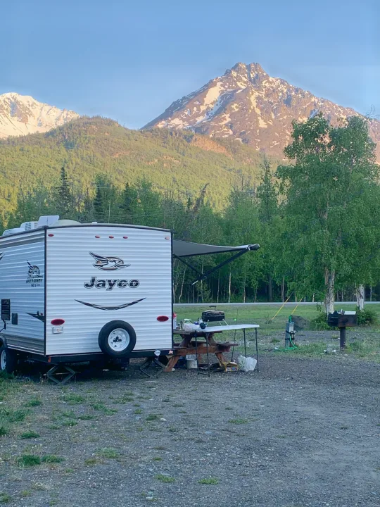 This RV rental is set up at a scenic campsite, showcasing a Jayco travel trailer against snow-capped mountains and green forests.. Jayco Jay Flight 2017