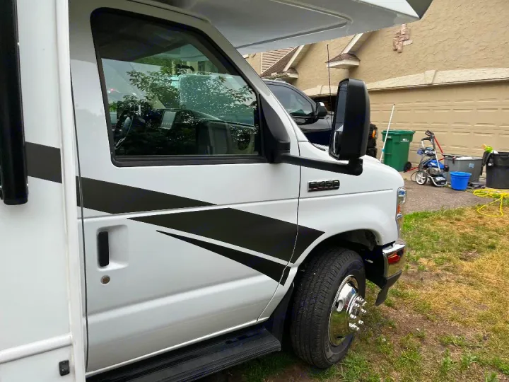 White RV with black graphic decals, large side mirrors, and shiny front wheel. This spacious rv rental is parked at a residence.. Jayco Redhawk 2021