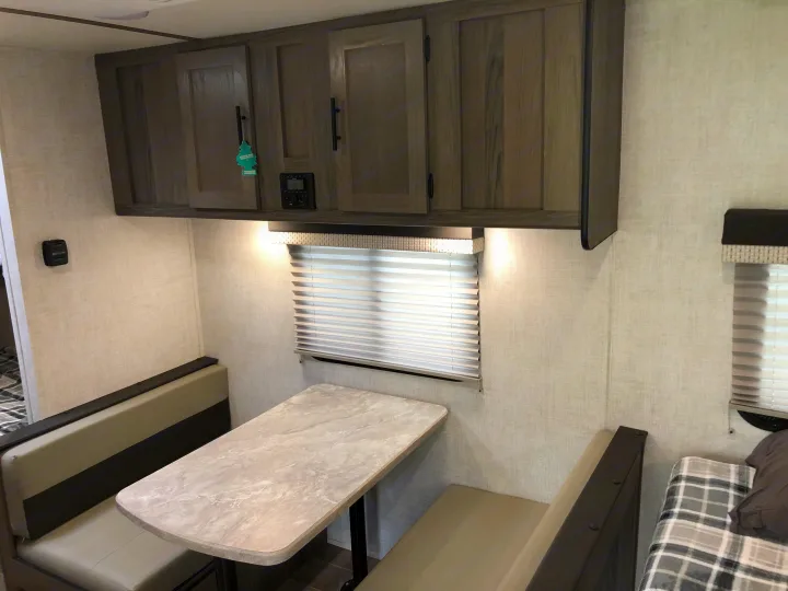 Cozy interior of an RV rental: a light-toned dinette with seating, a table, overhead cabinets, and a window with blinds.. Gulf Stream Ameri-Lite Super Lite 2021