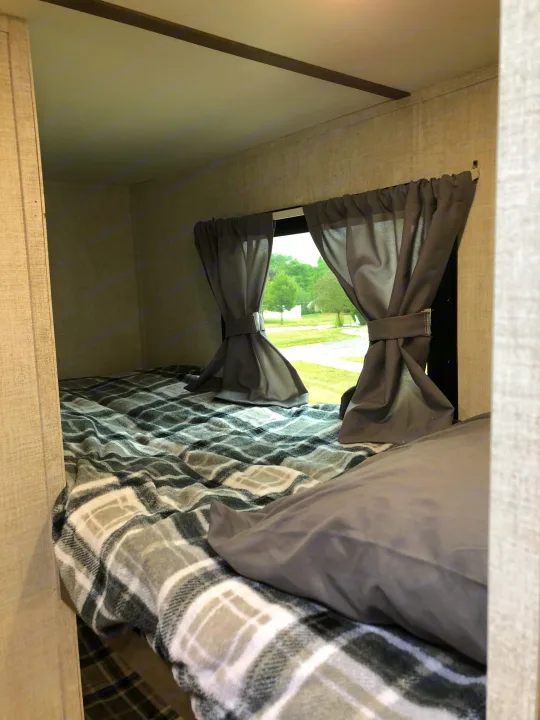 Cozy RV rental interior featuring a comfortable bed, window with tied-back grey curtains, and green outdoor view.. Gulf Stream Ameri-Lite Super Lite 2021