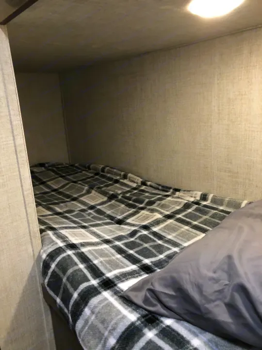 Cozy RV rental bunk bed with a grey plaid blanket and light-colored fabric walls. A bright light illuminates the snug space.. Gulf Stream Ameri-Lite Super Lite 2021